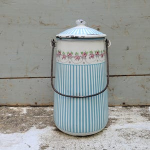 Puede incluir: Recipiente de esmalte vintage con tapa y asa. El recipiente presenta un patrón de rayas azules y blancas en el cuerpo, con una banda floral y una tapa a rayas azules y blancas. El asa de metal es oscura. El recipiente es cilíndrico.