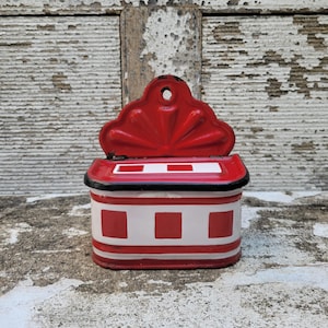 Op de afbeelding: Een vintage rode en witte emaille zoutdoos met een decoratief geschulpte deksel. De doos heeft een rechthoekige vorm met een rood-wit geblokt patroon op de voorkant en een zwarte rand. Het deksel heeft een klein gaatje om op te hangen.