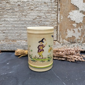 May include: A cylindrical ceramic mug with a painted illustration of a figure in a purple jacket and yellow pants. The mug has a cream-colored base with green and blue stripes near the bottom. The figure is holding a musical instrument.