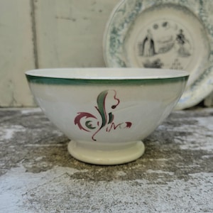 May include: Cream-colored ceramic bowl with a green rim and a decorative design in red and green. A matching plate with a faded green and white design is in the background. The bowl has a pedestal base.