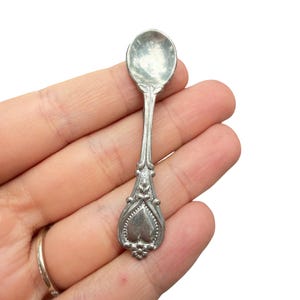 May include: A small silver-toned metal spoon featuring an ornate handle with a heart-shaped design at the base, decorative bead detailing along the edges, and a smooth, rounded bowl. The spoon has a polished finish and intricate relief patterns.