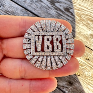 Peut inclure: Broche argentée avec un rectangle central portant les lettres "VBB". La broche circulaire présente un motif de rayons de soleil composé de petits cristaux transparents. La broche est tenue dans une main.