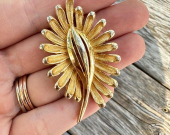 May include: A gold-tone vintage brooch in a floral design. The brooch features a central, textured element with a layered, open design, surrounded by radiating petals. The petals have a textured finish and are tipped with a silver-tone metal.
