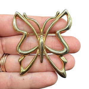 Puede incluir: Broche de mariposa dorada. El broche presenta un diseño calado con alas curvas y un cuerpo central. El alfiler se sujeta con un alfiler recto en la parte posterior. La mariposa es un motivo de joyería clásico.