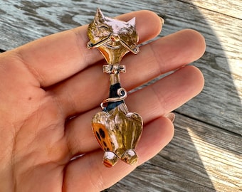May include: A gold-toned cat brooch with a smiling face, whiskers, and a black and gold bow tie. The cat's body has a heart shape with orange and black spots. The brooch is a vintage accessory.