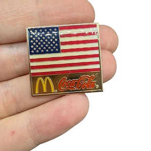 May include: A rectangular pin featuring the American flag design with red and white stripes and a blue field of stars. The bottom section displays the McDonald's golden arches logo and the Coca-Cola logo in red.