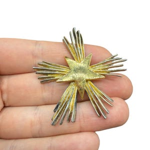 May include: A gold-toned starburst brooch with a central star and radiating rays. The brooch has a vintage aesthetic, with a mix of gold and silver tones. The starburst design is a classic, eye-catching accessory.