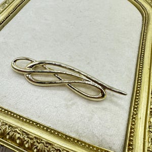 May include: A gold-toned brooch with an abstract design. The brooch features intertwined, flowing lines and curves, creating a sense of movement. The pin is set against a white background within a gold-colored frame.