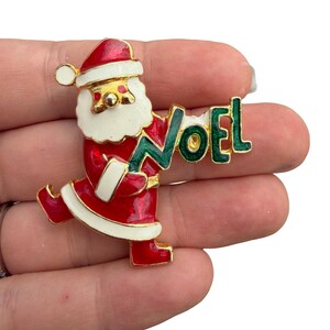 Puede incluir: Un broche de metal dorado con forma de Santa Claus con traje y gorro de esmalte rojo y blanco. Santa sostiene un letrero de esmalte verde que deletrea la palabra NOEL en letras mayúsculas. El diseño presenta esmalte brillante rojo, blanco y verde.