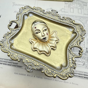May include: A gold-coloured metal tray with an ornate design. The tray features a raised, gold-coloured metal clown face with a red star and a red teardrop.