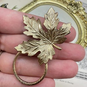 Pode incluir: Broche dourado em forma de folha com superfície texturizada e anel circular. O broche é projetado para se assemelhar a uma folha de bordo. O anel tem um design de corda torcida.