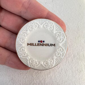 May include: White ceramic plate with a gold rim and intricate floral design. The center of the plate features the word "MILLENNIUM" in gold lettering with three small red and blue dots above it.