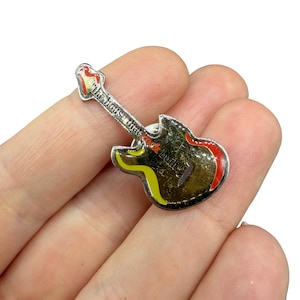 Op de afbeelding: Een zilverkleurige gitaarvormige pin met rode, gele en zwarte accenten. De pin heeft de tekst "the house that rock built" op de hals van de gitaar. De pin wordt in de hand van een persoon gehouden.