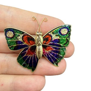 Puede incluir: Broche de mariposa dorada con alas de esmalte colorido. Las alas presentan una mezcla de acentos verdes, azules, rojos y dorados brillantes. El broche tiene un cuerpo y antenas detallados, y es un accesorio decorativo.