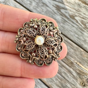 May include: A vintage silver-toned filigree brooch with a central pearl. The brooch has an intricate floral design with openwork details. The pearl is a creamy white color, and the metal has a slightly aged appearance.