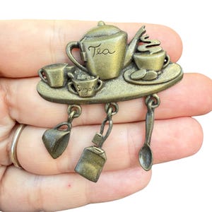 May include: A brass-toned metal brooch featuring a tea service scene with a teapot labeled Tea, a sugar bowl labeled Sugar, and a teacup. Three dangling charms hang from the base, including a small cup, a tea bag, and a spoon.