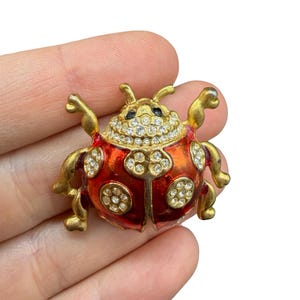 Op de afbeelding: Een goudkleurige lieveheersbeestjesbroche met een rode emaille behuizing en heldere kristallen accenten. De kop van het lieveheersbeestje is bedekt met kristallen en de vleugels hebben met kristal bezette cirkels. De broche heeft goudkleurige poten en antennes.