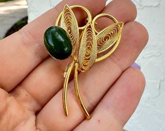 May include: A gold-toned brooch with a filigree design and a dark green oval stone. The brooch features two leaf-shaped elements and a central oval stone. The pin is a vintage style accessory.