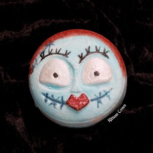 May include: A blue and red bath bomb shaped like Sally from The Nightmare Before Christmas. The bath bomb has a red heart-shaped mouth and white eyes with black pupils. The text "Kittens Coven" is printed on the bottom of the bath bomb.