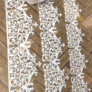 May include: White, ornate lace trim with a delicate floral design. The trim is arranged in vertical strips, showcasing intricate cut-out patterns. A soft pink rose rests at the top, adding a touch of color against the wooden background.