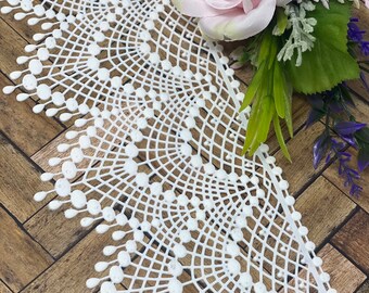 May include: White lace trim with a scalloped edge and a delicate pattern of circles and lines. The lace is shown on a wooden surface with a pink rose.