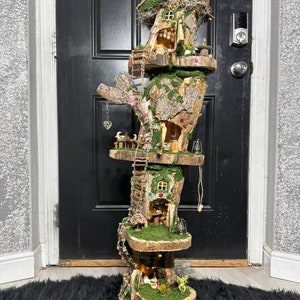 May include: A whimsical, multi-tiered fairy house sculpture. The structure features multiple levels with miniature houses, moss, and decorative elements. The color palette includes natural wood tones, green moss, and touches of color from the miniature details.