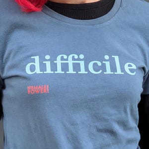 May include: A blue t-shirt with the word "difficile" printed in white on the front. The shirt also has the words "FEMALE POWER!" printed in pink on the front.