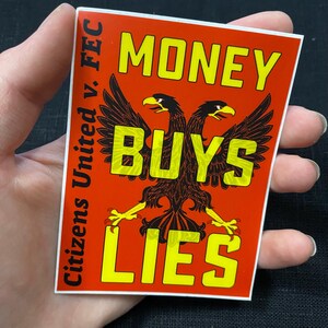 May include: A rectangular sticker with a red background and yellow text. The text reads "MONEY BUYS LIES" with "BUYS" in the center, and a two-headed eagle graphic. The words "Citizens United v. FEC" are on the left side.