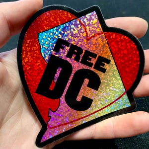 May include: A heart-shaped sticker with a red glitter border and a holographic center. The sticker has the words "FREE DC" in bold black letters. The sticker is held in a hand.