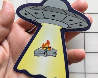 May include: A die-cut vinyl sticker featuring a gray UFO saucer with black circular accents. The UFO emits a bright yellow tractor beam downward, illuminating a small gray angular car that is engulfed in orange and red flames.