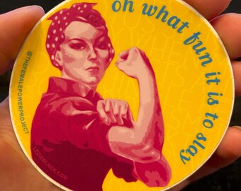 May include: A circular sticker with a yellow background featuring a red-toned illustration of a person flexing their arm while wearing a polka-dot headscarf. The text reads "oh what fun it is to slay", "@THEFEMALEPOWERPROJECT", and "LEDABLACK.COM".