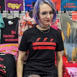 May include: A person wearing a black t-shirt with a red horizontal stripe and three red rats printed on the stripe. The shirt has short sleeves and a crew neck. The person has blue hair and is wearing a silver chain necklace with a black studded choker.