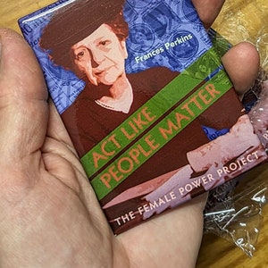 May include: A blue and green button with the text "Act Like People Matter" and "The Female Power Project" with a portrait of Frances Perkins.
