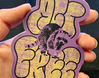 May include: A die-cut sticker featuring a purple and yellow textured background with the text "ACT FREE" in a bold, rounded font. A black and purple illustration of a raccoon is centered over the text. The text "ACTFREE.STUDIO" is printed in small letters at the bottom.
