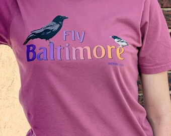 May include: A mauve short-sleeve t-shirt featuring a graphic of a black crow and a smaller black and white bird. The text reads Fly Baltimore in a multi-colored gradient font, with the smaller text ACTRE below the word Baltimore.