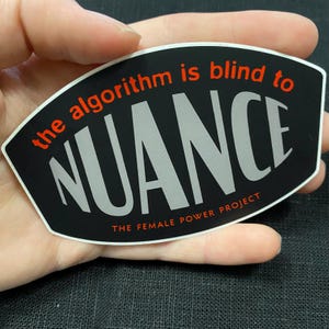 May include: A black sticker with the words "the algorithm is blind to NUANCE" in red and silver lettering. The text "THE FEMALE POWER PROJECT" is at the bottom. The sticker is held in a hand.