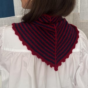 May include: A triangular crochet neck warmer featuring alternating dark blue and deep red stripes. The accessory has a textured knit pattern and is finished with a scalloped red crochet border along the entire outer edge of the triangular shape.