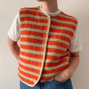 Könnte beinhalten: Eine gestrickte Weste mit horizontalen orangefarbenen und hellbeigen Streifen. Die Weste hat einen runden Ausschnitt und wird vorne mit beigen Knöpfen geschlossen. Die Weste wird über einem weißen T-Shirt und einer blauen Jeans getragen.