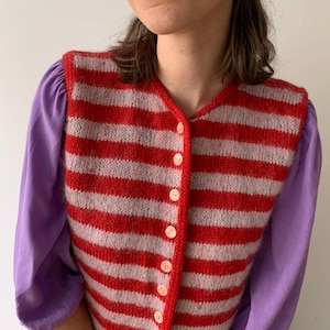 Könnte beinhalten: Eine rot-weiß gestreifte Strickweste mit V-Ausschnitt und Knopfverschluss. Die Weste wird über einem lila Langarm-Oberteil getragen. Die Knöpfe sind hellrosa.