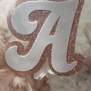May include: A white letter A with rose gold glitter trim. The letter is surrounded by white paper flowers and pink feathers.