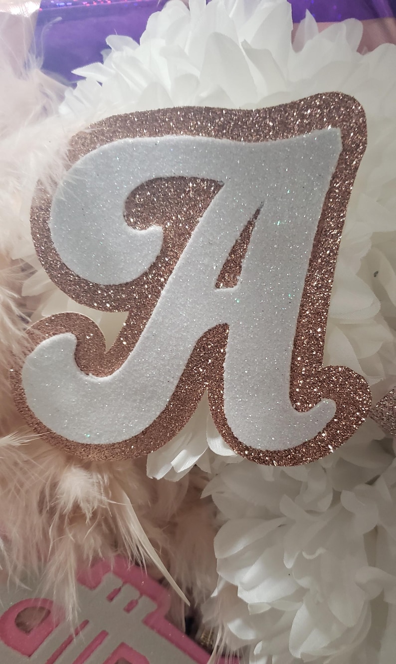 May include: A white letter A with rose gold glitter trim. The letter is surrounded by white paper flowers and pink feathers.