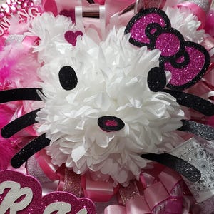 May include: A white, fluffy, Hello Kitty-inspired flower with black glitter accents and a pink glitter bow. The flower is surrounded by pink and white ribbons and feathers.