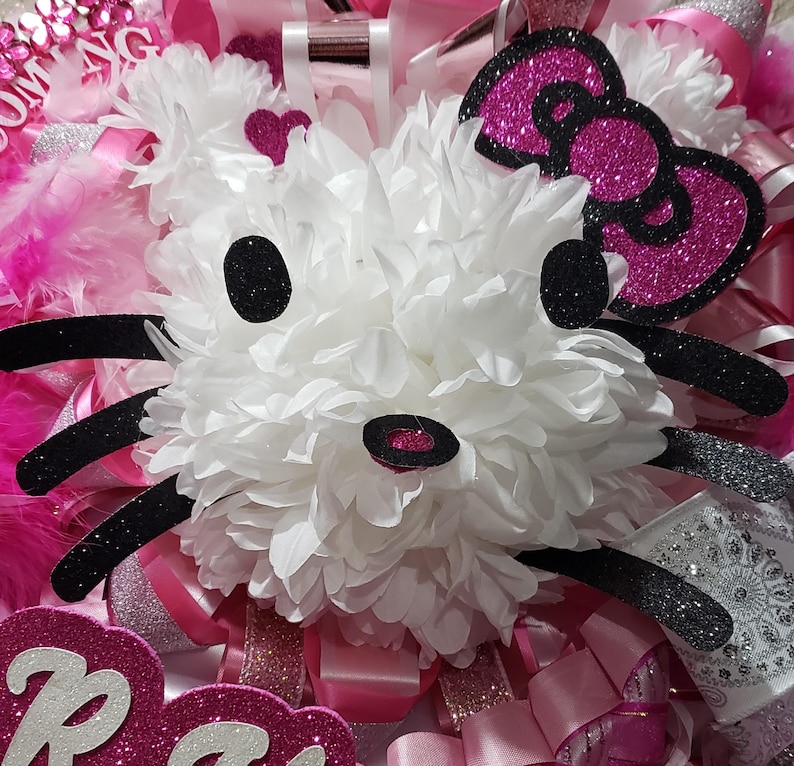 May include: A white, fluffy, Hello Kitty-inspired flower with black glitter accents and a pink glitter bow. The flower is surrounded by pink and white ribbons and feathers.