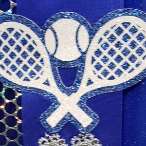 May include: Two white tennis rackets with blue glitter, crossed over each other, with a white tennis ball in the center. The rackets and ball are on a blue background.
