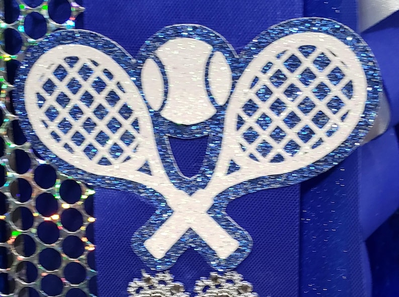 May include: Two white tennis rackets with blue glitter, crossed over each other, with a white tennis ball in the center. The rackets and ball are on a blue background.