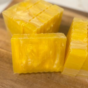 May include: Close-up of several bars of yellow soap. The soap has a translucent quality and a shimmering, golden appearance. The bars are cut into rectangular shapes, with some sliced into thinner pieces. The soap is arranged on a wooden surface.