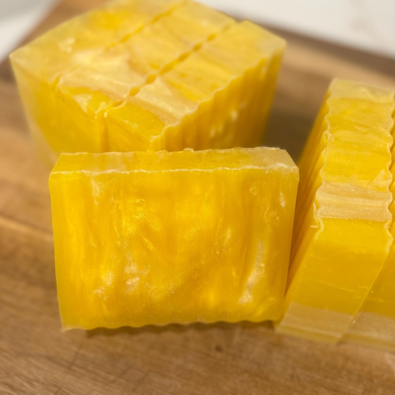 May include: Close-up of several bars of yellow soap. The soap has a translucent quality and a shimmering, golden appearance. The bars are cut into rectangular shapes, with some sliced into thinner pieces. The soap is arranged on a wooden surface.
