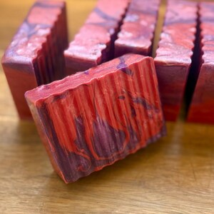 Pu&ograve; includere: Diverse saponette rettangolari caratterizzate da un motivo marmorizzato con vortici di rosso intenso, arancione brillante e viola scuro. Ogni saponetta presenta un bordo superiore testurizzato e scanalato e una superficie frontale liscia.