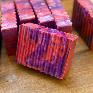 Pu&ograve; includere: Una collezione di saponette rettangolari caratterizzate da un motivo marmorizzato con vortici di rosso vivace, viola intenso e bianco. Ogni saponetta presenta una superficie testurizzata e scanalata sul lato anteriore, con colori miscelati.