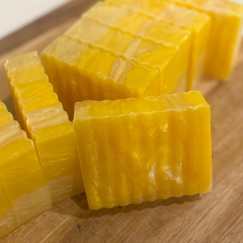 May include: Close-up of several bars of yellow soap, cut into rectangular shapes. The soap has a translucent quality with a slightly textured surface. The bars are arranged on a wooden cutting board, showcasing the handmade nature of the product.